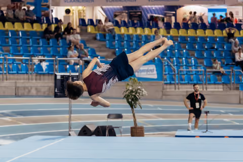Young gymnast soaring through a powerful backflip, body fully extended, in a blue indoor sports arena.