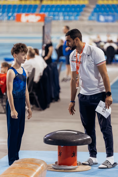 A young male gymnast listens attentively as his coach leans in to give instructions beside the vault apparatus.