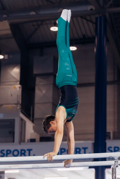 Young male gymnast performs a precise handstand on parallel bars, wearing teal and black attire, in an indoor gymnasium.