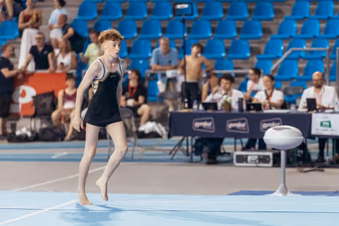 Young gymnast with red hair lands barefoot on floor mat, focused expression, judges table visible in background.