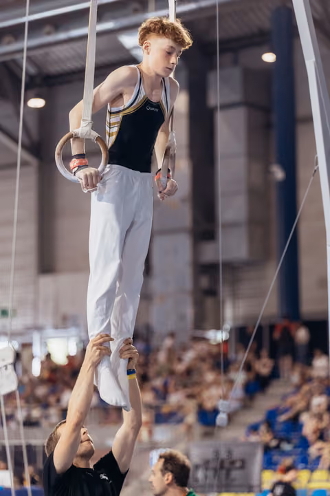 Young male gymnast grips still rings with focused concentration while coach steadies his legs in packed arena.