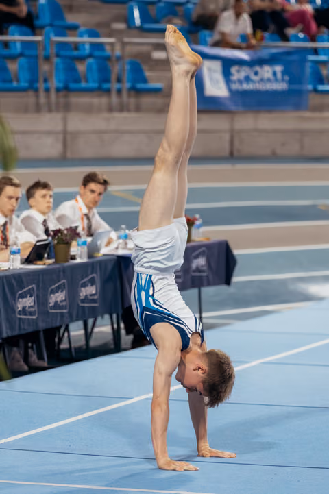 Young male gymnast performing a perfect handstand on the floor, legs fully extended upward, at a competitive gymnastics event.