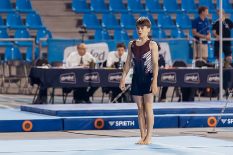 Young male gymnast stands barefoot on the floor exercise mat, composed and focused, at a Gymfed competition.