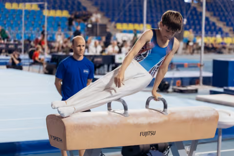 Young male gymnast performs on pommel horse with intense focus while coach observes attentively in background.