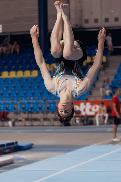 Male gymnast captured mid-backflip, fully inverted above the floor exercise mat in a competition arena.