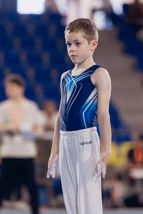 Young male gymnast stands composed and focused, wearing a navy blue leotard and white pants, chalked hands at sides, in a gymnasium.