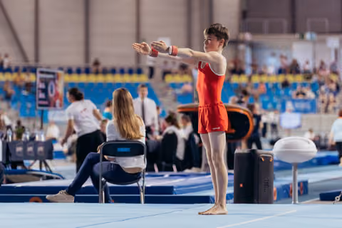 Young male gymnast in red leotard stands poised on floor mat, arms extended forward, focused expression before routine.