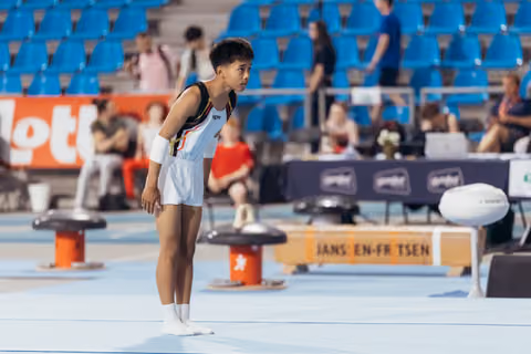 Young male gymnast in Belgian leotard stands focused on the floor, leaning forward in concentration before his routine.