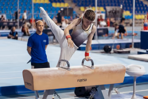 Young male gymnast performs scissors on pommel horse, concentrated expression, coach watching attentively in background.