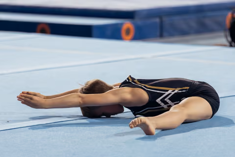 Young gymnast in black leotard stretches forward on blue floor mat, arms extended, head down in focused warm-up.