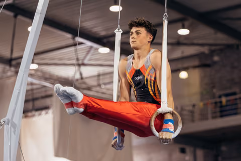 Young male gymnast holds an L-sit on still rings, wearing red pants and agiva kit, focused expression in indoor gym.