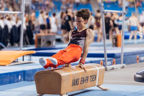 Young male gymnast performs focused pommel horse routine, legs raised, wearing red pants in a busy sports hall.