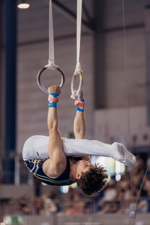 Young male gymnast performs an inverted hold on still rings, showing strength and concentration during a competition.
