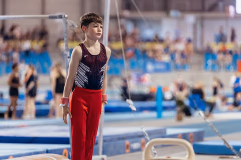 Young male gymnast stands composed and focused near the parallel bars, wearing red trousers and a dark leotard.