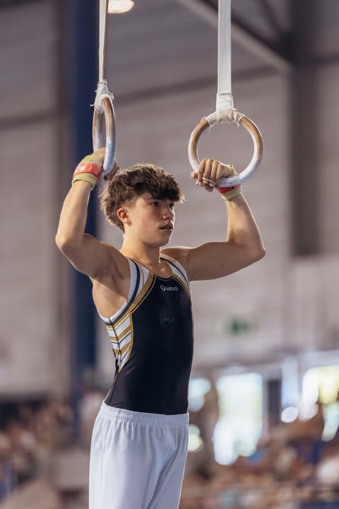 Young male gymnast grips still rings with focused intensity, wearing a black Quatro leotard in a gymnastics arena.