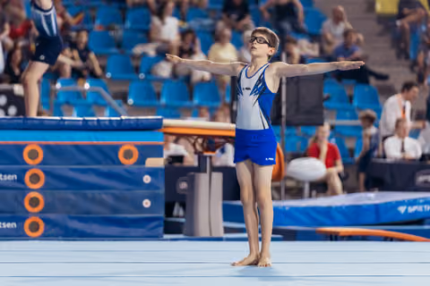 Young male gymnast stands barefoot with arms outstretched, composed and focused, on the floor exercise mat in a busy arena.