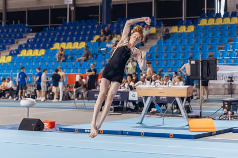 Young male gymnast leaps with arms raised and fists clenched in triumph during floor exercise at indoor arena.