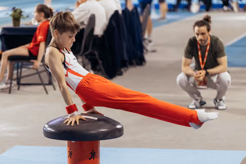 Focused young male gymnast performs a straddle hold on a mushroom while a coach watches attentively in the background.
