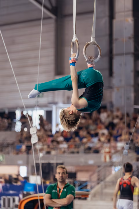 Young male gymnast hangs inverted on still rings in green uniform, coach watching intently below in packed arena.