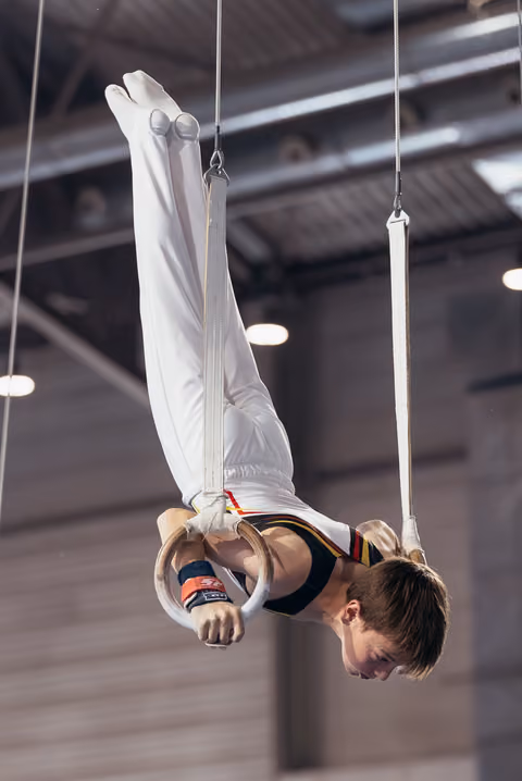 Young male gymnast performs an inverted iron cross on still rings, face tense with concentration, in an indoor gymnasium.