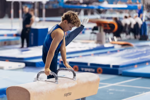 Young male gymnast, focused and composed, grips pommel horse handles with chalked hands in a blue gymnastics hall.