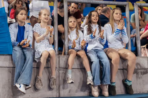Five young girls sitting on stadium wall, clapping enthusiastically while watching a sports event, smiling and engaged.