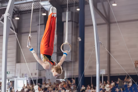 Young male gymnast performs an inverted hold on still rings, body fully extended upside down, wearing red and black leotard.