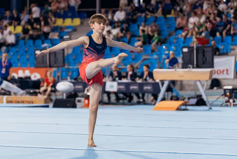 Young male gymnast balances on one leg with arms outstretched during a floor exercise routine at an indoor arena.