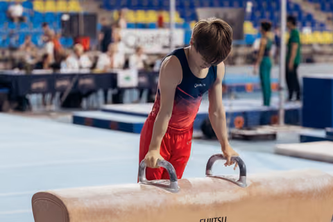 Young male gymnast grips pommel horse handles, leaning forward with intense concentration during a gymnastics competition.