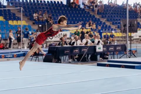 Young male gymnast in red leotard soars across the floor exercise mat, arms extended forward, body fully stretched mid-flight.