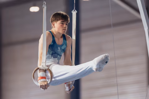 Young male gymnast performs on still rings, legs raised, expression focused and determined in an indoor gymnasium.