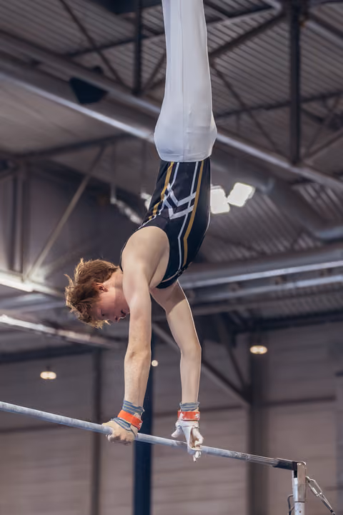 Red-haired male gymnast performs an inverted hold on the high bar, gripping with wrist guards, intense concentration in a gymnastics hall.