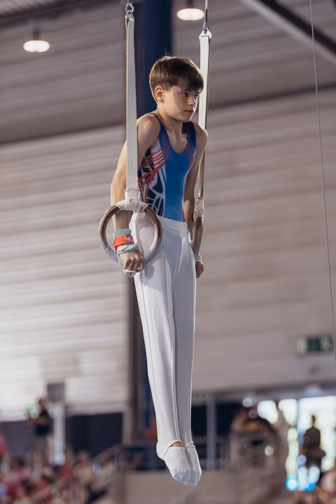Focused young male gymnast holding still rings in iron cross position, wearing blue leotard and white pants in gymnasium.