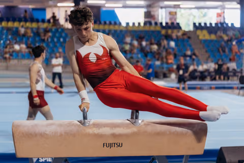 Young male gymnast performs on pommel horse, body parallel, intense focus. Fujitsu-branded apparatus in arena.