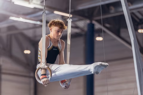 Young male gymnast holds an L-sit on still rings, focused expression, wearing black leo and white pants in a gymnastics hall.