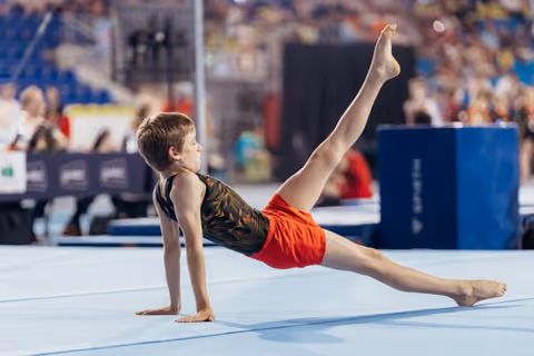 Young gymnast performs a floor exercise, raising one leg high with focused determination in a competition arena.