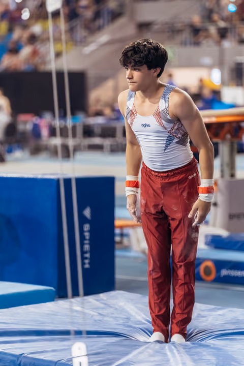Young male gymnast stands composed on the mat, chalked hands relaxed, gazing intently aside in a busy competition arena.