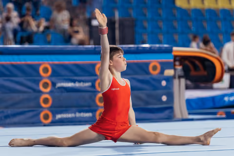 Young gymnast performs a side split on the floor with one arm raised high, focused expression in a blue arena.