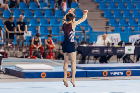 Young male gymnast leaps with arms raised triumphantly on the floor exercise mat during a gymnastics competition.