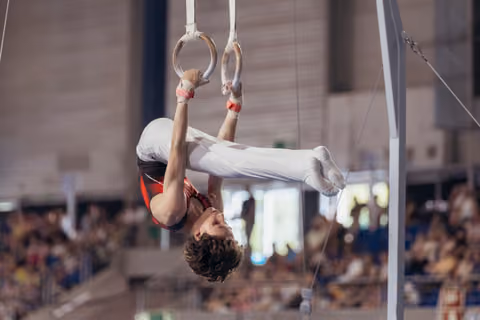 Young male gymnast hangs inverted on still rings, body curved with intense concentration during a packed arena competition.