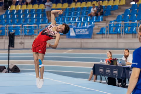 Young male gymnast in red leotard soars mid-air with arched back during floor exercise at indoor sports arena