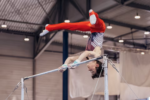 Young gymnast in red performs an inverted release on the horizontal bar, smiling mid-skill in an indoor gymnasium.