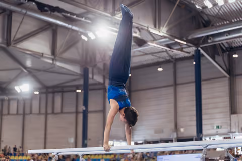Young male gymnast performs a perfect handstand on parallel bars, body fully extended, in a packed sports arena.
