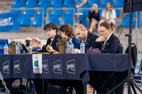 Four judges in dark blazers sit focused at a Gymfed judging table, reviewing scores during a gymnastics competition.