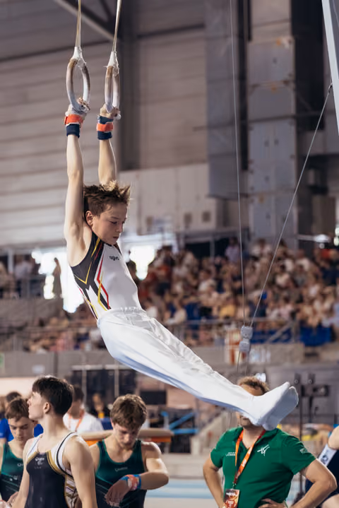 Young male gymnast performs an L-sit on still rings, focused expression, white uniform, crowd watching in background.