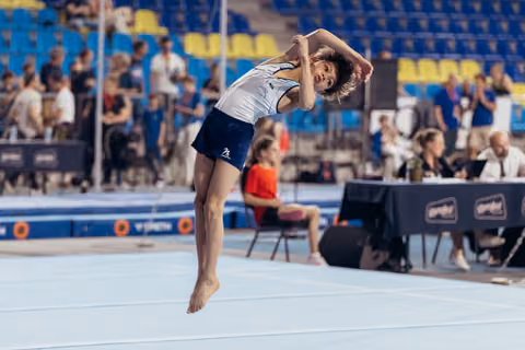 Young gymnast leaps mid-air on floor exercise, body arched gracefully, arms framing head, focused expression during competition.
