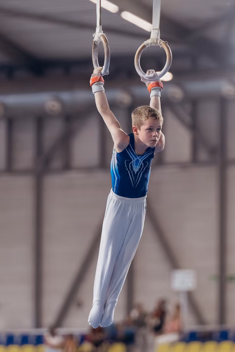 Focused young male gymnast hangs from still rings, legs together, intense concentration, in a gymnasium during competition.