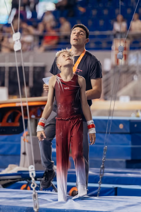 A coach stands behind a young gymnast in red, both looking upward with focus and guidance on a trampoline.
