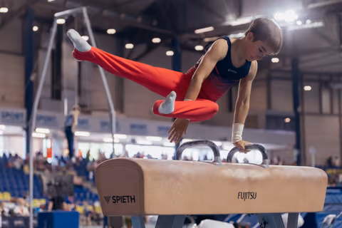 Young male gymnast performs scissor move on pommel horse, legs split wide, intense focus, in a packed competition arena.