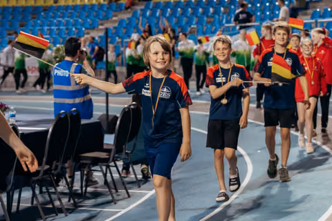 Smiling children in navy uniforms wave Belgian flags proudly during a victory parade on an indoor athletics track.